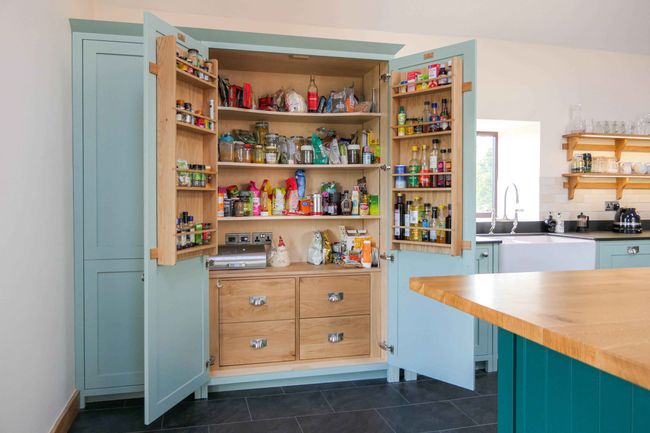 Open light blue pantry with wooden shelves filled with various food items and spice rack on either side. Shaker kitchen background with a wooden countertop and sink.