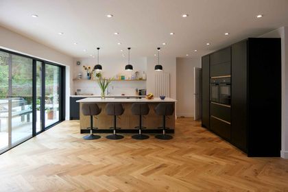 Modern kitchen with a central island, four bar stools, black cabinetry, pendant lights, and large windows overlooking greenery.