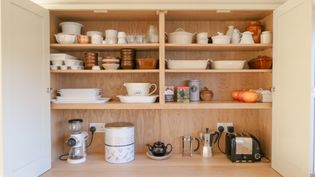 A wooden kitchen cupboard filled with various types of dishes, including plates, bowls, and mugs, as well as kitchen appliances like a coffee maker and toaster.