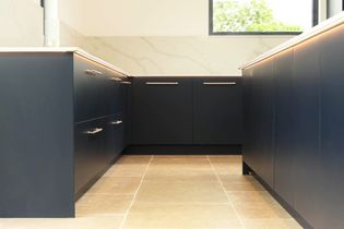 Modern kitchen with dark blue cabinets, silver handles, and beige tiled floor. Marble effect backsplash and natural light from a window create a sleek, airy feel.