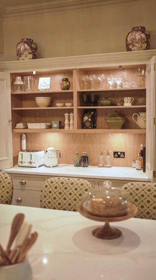 A well-organised kitchen dresser with shelves displaying various dishware, glassware, and kitchen appliances, including a toaster and a kettle, with decorative items on top.