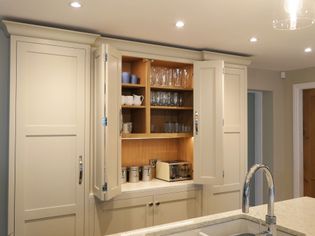 Shaker kitchen with cream cabinets, open to reveal shelves with glasses and cups. A toaster and canisters are on the countertop, under soft lighting.