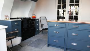 Shaker kitchen with sleek blue cabinets, a black range, and a copper countertop on the island. A red kettle and two chairs add warmth to the clean, inviting space.