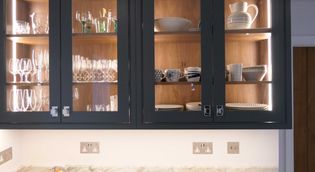Illuminated kitchen cabinets with glass doors display various glassware and ceramic dishes. The modern and organized layout conveys elegance and order.