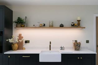 Shaker kitchen with black cabinets, white countertops, and a farmhouse sink. A wooden shelf above holds decorative items including plants and candles.