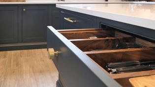 Open kitchen drawer with dark wood organizer, holding neatly arranged silverware. Modern design with brass handles and light wood floor in background.