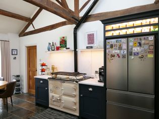 Rustic kitchen with exposed wood beams, slate floor, and cream vintage stove. A fridge with magnets stands nearby. Warm, cozy atmosphere.