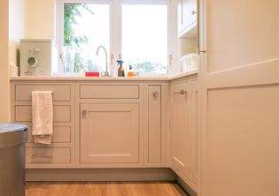 Neutral kitchen with a sink beneath two windows, surrounding Shaker cabinets, a hanging towel, and countertop supplies.